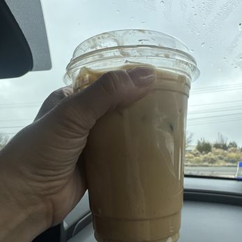 Juniper - Pumpkin spice latte iced with oat milk. - A person holds a plastic cup of iced coffee with oat milk inside a car, with a rainy windshield in the background. - Coffee & Tea Near Me - Farmington, New Mexico