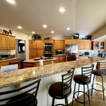 Jacob's House - Kitchen/dining area, with designated cabinets and refrigerator shelves for each family. - The image shows a kitchen with wooden cabinetry, including upper and lower cabinets, and a large granite countertop island. The breakfast bar has black metal stools with cushioned seats. The kitchen contains stainless steel appliances, including a refrigerator, oven, microwave, and toaster. The room is illuminated by multiple ceiling lights, and decorative items are placed on top of the cabinets. The flooring appears to be hardwood, and the space is open and organized. - Community Service/Non-Profit Near Me - Temecula, California