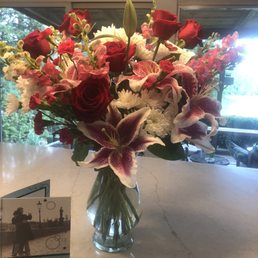 Anniversary floral arrangement - A bouquet of red roses, pink lilies, and white daisies arranged in a glass vase on a marble table near a window.