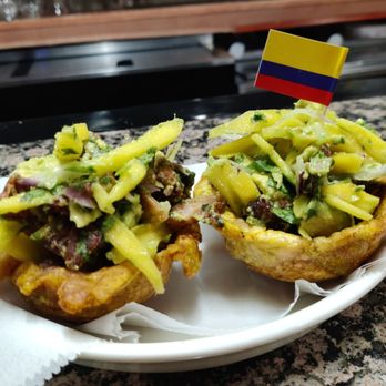 Song & Sazon - Ceviche de chicarron - Two Colombian-style chicharrón cups filled with a vegetable salad, including sliced vegetables and herbs, topped with a small Colombian flag, placed on a white plate on a granite countertop. The setting appears to be a restaurant with a granite surface and a wooden background. - Colombian Near Me - Silver Spring, Maryland