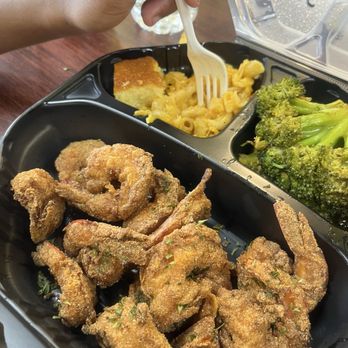 Oooh We It Is - A black plastic takeout container contains fried shrimp seasoned with herbs, macaroni and cheese with a creamy sauce, steamed broccoli, and a piece of cornbread. A child's hand is holding a fork, picking at the macaroni and cheese. In the background, there is a wooden table, a cup, and a bottle of fruit punch. The food appears to be from a soul food or Southern-style restaurant. - American Near Me - Chicago, Illinois