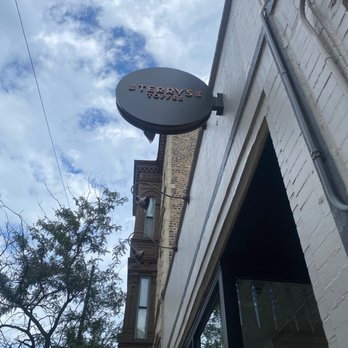 Terry's Toffee - The image shows a circular sign for Terry's Toffee mounted on the exterior wall of a building. The sign is black with red and white lettering. The building has a white brick facade, and part of a large window is visible at the bottom of the image. The background features a partly cloudy sky with some trees visible on the left side of the image. - Candy Stores Near Me - highland park, Illinois
