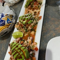 Trio de Ceviche - Three ceviche portions with diced seafood, vegetables, and avocado slices are arranged on a rectangular white plate, garnished with sauce and a lime wedge.