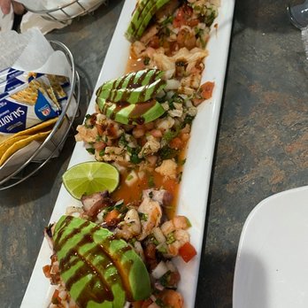 La Costa Bora - Trio de Ceviche - Three ceviche portions with diced seafood, vegetables, and avocado slices are arranged on a rectangular white plate, garnished with sauce and a lime wedge. - Seafood Near Me - Belvidere, Illinois