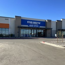 The image depicts the exterior of a Five Below store located in a shopping center, with a prominent blue and white sign reading 'Five Below.' The storefront features large glass windows and a glass entrance door. The surrounding area includes a parking lot with a few cars and small landscaped islands with young trees and gravel. The sky above is clear and blue, indicating a sunny day.