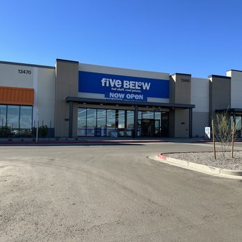 Five Below - The image depicts the exterior of a Five Below store located in a shopping center, with a prominent blue and white sign reading 'Five Below.' The storefront features large glass windows and a glass entrance door. The surrounding area includes a parking lot with a few cars and small landscaped islands with young trees and gravel. The sky above is clear and blue, indicating a sunny day. - Discount Store Near Me - Surprise, Arizona