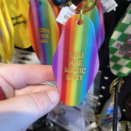 Fun keychains! - The image shows a rainbow-colored, diamond-shaped keychain with gold embossed lettering that says "YOU ARE MAGIC BABY." The keychain is attached to a gold keyring and is held by a person's hand. In the background, there are other similar rainbow-colored keychains with different messages, hanging among various other keychains and accessories in a retail display setting.