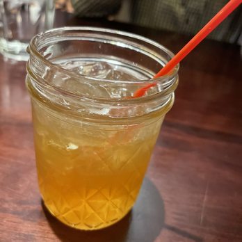 Primo - Whisky in a jar drink special - A mason jar filled with a yellowish drink and ice cubes is positioned on a wooden table. An orange straw is inserted into the jar. The background shows a blurred restaurant interior with glasses and a person wearing a checkered shirt. The setting suggests a casual dining environment, possibly at Primo in New Castle, IN, as indicated by the caption. - Italian Near Me - New Castle, Indiana