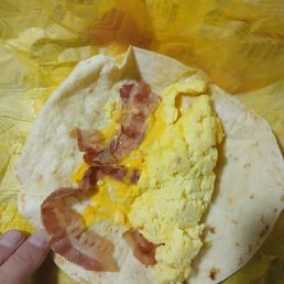This is considered well done! - A breakfast burrito with a flour tortilla is partially unwrapped, showing scrambled eggs, crispy bacon strips, and melted cheese inside. The burrito is placed on yellow wax paper, and a person's fingers are holding the edge of the tortilla.