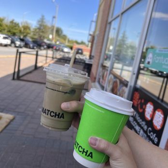 Matcha Matcha - Two individuals are holding matcha drinks, one in a clear plastic cup with a lid and straw, and the other in a green paper cup with a lid. They are standing outside a cafe with large glass windows, and the background shows parked cars, a sidewalk, and a bright, clear sky. The scene suggests a casual outdoor coffee or tea outing. - Coffee & Tea Near Me - Markham, Ontario