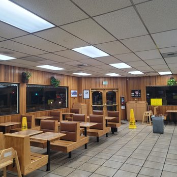 The Hat - ready to close - The image shows an indoor dining area of a restaurant with wooden wall paneling and ceiling tiles. There are multiple tables and chairs arranged throughout the space, with some chairs upholstered in brown material. Potted plants are placed on the window ledges and wall shelves. The floor is tiled, and there are yellow caution signs on the floor. Double glass doors with an exit sign above are visible at the back, and large windows line the walls, showing a nighttime scene outside. - American Near Me - Pasadena, California