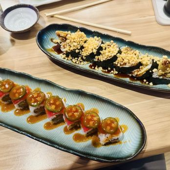Sushi Bene - Two elongated plates of sushi with different toppings and sauces are on a wooden table, accompanied by small bowls and drinks. - Japanese Near Me - Prosper, Texas