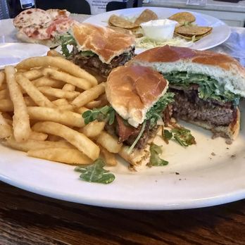 The Quarry Thornton - The Quarry burger - A burger with a beef patty, lettuce, tomato, and greens, served on a toasted bun, cut in half to reveal the ingredients. The plate also contains a portion of French fries. In the background, there are additional food items, including a pizza and nachos, on a table at a restaurant setting. - Bars Near Me - Thornton, Illinois