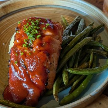 Subourbon - Salmon - A ceramic plate holds a serving of meat topped with a red sauce and garnished with chopped green herbs. Next to the meat, there is a portion of cooked green beans. The plate is placed on a wooden table, with other dishes and drinks visible in the background. - Southern Near Me - Columbus, Ohio