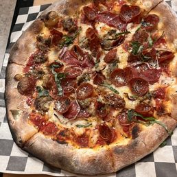 A pizza with a slightly charred, rustic crust features toppings including pepperoni, sausage, and fresh herbs. The pizza is placed on checkered paper on a wooden table, showcasing a variety of meat toppings and melted cheese.