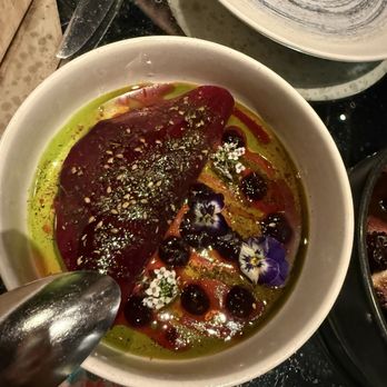 ilili DC - Beetroot - A ceramic bowl holds a piece of cooked meat, garnished with edible flowers, herbs, and berries. The dish is surrounded by a green oil-based sauce and a red sauce, with visible seasoning and oil glistening on the surface. The presentation includes floral and berry elements, indicating a gourmet or fine dining preparation. - Lebanese Near Me - Washington, DC, District of Columbia