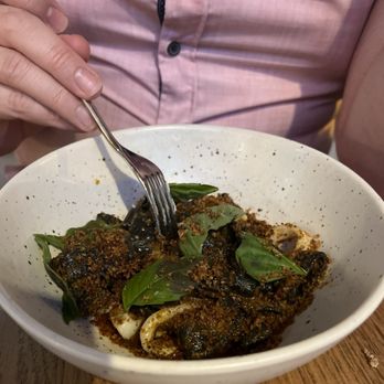 Wildweed - Chorizo Squid Ink Pasta - A person in a pink shirt is holding a fork over a bowl of pasta with basil leaves and a dark sauce, likely at a restaurant. - New American Near Me - Cincinnati, Ohio