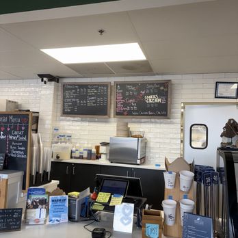 Maple Leaf Coffee House - The image shows a coffee shop counter with a white surface, a cash register, and various supplies including cups and utensils. Behind the counter, there are menu boards with drink options and prices, a stainless steel appliance, and a white tiled wall. To the right, there are stacks of cups and a small wooden block labeled 'TIPS!'. The setting appears to be a casual coffee shop environment. - Cafes Near Me - St. Charles, Illinois