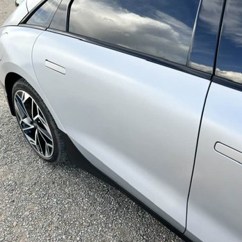 Body Master - The image depicts a silver electric vehicle parked on a gravel surface. The car's side panel, rear wheel, and part of the window are visible. The windows reflect a cloudy sky, indicating an outdoor setting. The vehicle features modern design elements, including sleek lines and stylish alloy wheels, and is positioned in an open area with no other objects or vehicles in immediate view. - Body Shops Near Me - Albuquerque, New Mexico