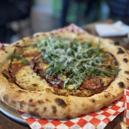 Figgy Piggy - A wood-fired pizza featuring a slightly charred, golden-brown crust, topped with fresh arugula, slices of prosciutto, and grated cheese. The pizza is placed on a tray lined with red and white checkered paper, with a blurred background indicating a restaurant setting.