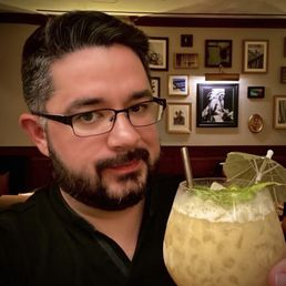 YOU'RE THE SUNFLOWER  Cacao Nib Infused White Rum, Coconut Rice Milk, Passionfruit, & Lime! - A man with dark hair, glasses, and a beard is taking a selfie in a restaurant or bar setting. He is holding a large cocktail glass filled with a yellowish drink, garnished with a small umbrella, lime slices, and a straw. The background features a wall decorated with various framed pictures and artwork, creating a cozy and artistic atmosphere.