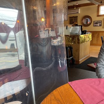 Brickhouse Cafe - Plexiglass divider - The image depicts the interior of a restaurant with a clear plastic divider placed on a wooden table with a red placemat. Behind the divider, a person is seated, partially visible through the reflection and transparency of the plastic. The background includes a counter with a gold tinsel decoration, a mirror, framed photos, and an air conditioning unit. The ceiling features exposed ductwork and warm lighting, creating a cozy atmosphere. - Italian Near Me - Dedham, Massachusetts