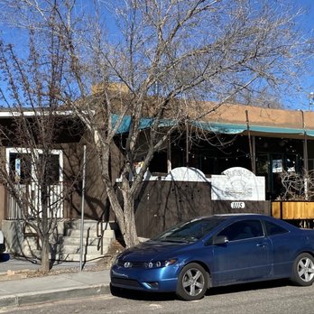 Tune-Up Cafe - Outside of cafe - The photo depicts the exterior of Tune-Up Cafe, a restaurant with a brown facade and a sign displaying its name. There are parked vehicles, including a blue car and a white SUV, along the street. Leafless trees are present in front of the cafe, and the sky is clear and blue, indicating a sunny day. - New Mexican Cuisine Near Me - Santa Fe, New Mexico