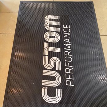 Custom Performance - A rectangular black rubber mat with a textured surface features the words "CUSTOM PERFORMANCE" printed in white. The mat is positioned on a beige tiled floor, with visible grout lines and some minor surface imperfections. The image is taken from an overhead angle, showing the entire mat and part of the surrounding floor. - Physical Therapy Near Me - New York, New York