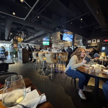 Kitchen and Rail - A woman sits at a round table in a restaurant, surrounded by other diners and a bar area, with a glass of white wine in the foreground. - Bars Near Me - Eagan, Minnesota