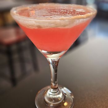 Double Ts Tavern - A pink-colored cocktail in a martini glass is placed on a dark table, with a blurred background of a bar or restaurant setting. - Bars Near Me - Marlborough, Massachusetts