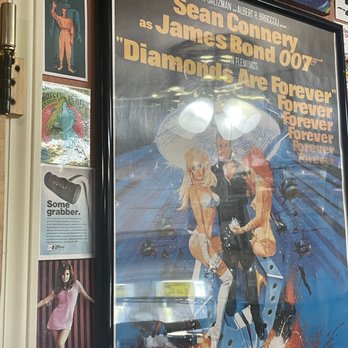 Grumpy's - The image shows a framed poster for the James Bond film 'Diamonds Are Forever,' featuring artwork of James Bond with two women, set against a stylized background. The poster includes text with the names of actors and the film title. To the left of the poster, there are smaller images and a sign about a product called 'HURST.' The setting appears to be an indoor space with various decorations and posters on the wall. - American Near Me - Gravette, Arkansas