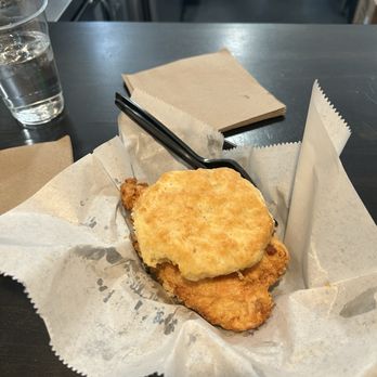The West Egg - Pimento cheese chicken sandwich - A fried chicken sandwich topped with a biscuit is placed on parchment paper on a dark table, with a glass of water and napkins nearby. - Coffee & Tea Near Me - St. Augustine, Florida