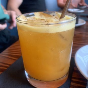 Corner Bar and Kitchen - Bourbon sour - A transparent glass holds a bourbon sour cocktail with ice cubes, a straw, and a frothy yellowish top layer. The drink is set on a black napkin on a wooden table. In the background, there are people seated, with one person wearing a dark shirt and another in a pink top, suggesting a social dining or drinking setting. - Cocktail Bars Near Me - Rangeley, Maine