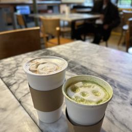 Latte and matcha - Two cups of drinks, a matcha latte and a coffee, are placed on a marble table in a cafe with wooden chairs and large windows.