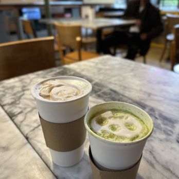 Cafe Milano - Latte and matcha - Two cups of drinks, a matcha latte and a coffee, are placed on a marble table in a cafe with wooden chairs and large windows. - Cafes Near Me - Berkeley, California