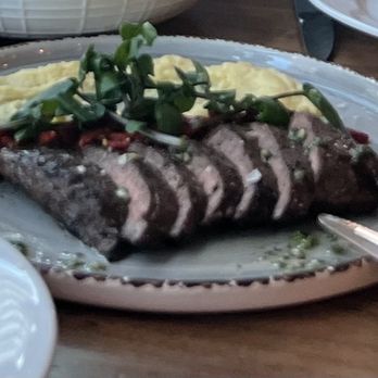Marina - Wagyu Flat Iron - Sliced beef steak with a pink center, topped with green garnish, served alongside mashed potatoes and a red sauce on a ceramic plate. The dish is placed on a wooden table with other plates partially visible around it. - Italian Near Me - Pasadena, California