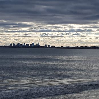 Dockside Pub - View of ocean and Boston skyline across the street. - The image depicts a body of water, likely the ocean, with gentle waves near the shore. In the distance, a city skyline with multiple high-rise buildings is visible across the water. The sky is filled with layered clouds, creating a moody atmosphere. The scene is viewed from a shoreline or beach area, with a rocky outcrop on the right side of the image. - Bars Near Me - Swampscott, Massachusetts