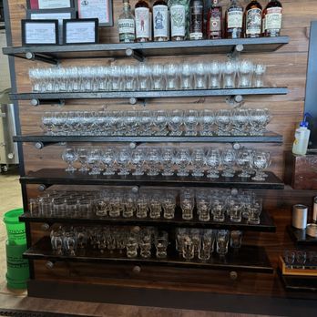 Analytical Brewing - Multiple black shelves are mounted on a wooden wall, displaying an array of glassware including wine glasses, shot glasses, and beer glasses. Bottles of liquor are placed on the top shelf. To the right, a person’s arm is visible, along with a small bottle of sanitizer and some cleaning cloths. The setting appears to be a bar, brewery, or tasting room with a focus on drinkware and alcohol bottles. - Beer Bar Near Me - Lexington, Illinois