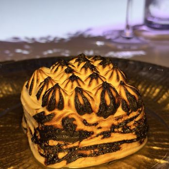 Elysium Immersive Dining - Tiramisu - A tiramisu dessert with a layered, textured surface showing dark and light swirls, presented on a glass plate. The background includes a blurred table setting with wine glasses and a glass of water, indicating a dining environment. - Modern European Near Me - Lighthouse Point, Florida