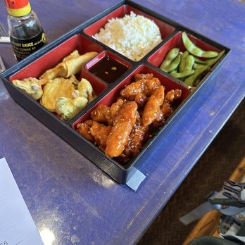Yu Garden - General Tso Chicken Bento Box - A Japanese-style bento box contains rice, glazed chicken, edamame, fried items, and soy sauce, placed on a blue table. - Chinese Near Me - Red River, New Mexico