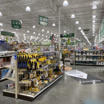 Menards - The image shows the interior of a large hardware store with high ceilings and bright lighting. Shelves are filled with various tools, adhesives, and hardware supplies, organized in sections. Signage hangs from the ceiling indicating different product categories. The floor is made of polished concrete, and there are promotional signs and price tags visible throughout the aisle. - Hardware Stores Near Me - Indianapolis, Indiana