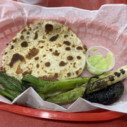 Cheese Quesadilla - A food tray contains a cheese quesadilla with browned spots, a small cup of green chili sauce, grilled green vegetables, and a roasted pepper. The items are arranged on pink paper inside a red plastic tray, typical of casual dining or takeout settings.