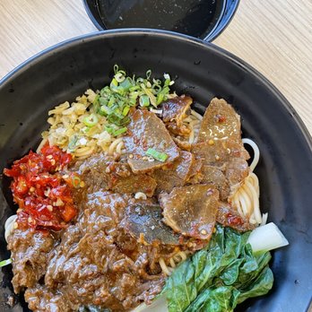 Jin’s Mi-Fen - A black bowl contains Asian noodles topped with beef chunks, chopped green onions, and vegetables including leafy greens and diced red chili. A small portion of chopped garlic or ginger is also visible. A black cup filled with a dark beverage, possibly tea or soda, is placed behind the bowl on a light wooden surface. - Chinese Near Me - Markham, Ontario