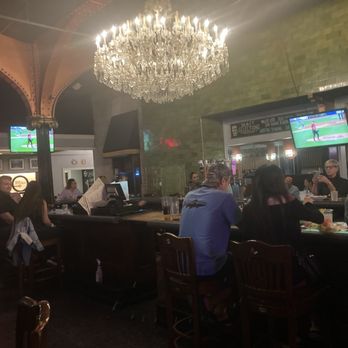 Trax - Funky bar - The image shows a bar with a large, ornate chandelier hanging from the ceiling. Patrons are seated at the bar, watching sports on two TV screens mounted on the wall. The bar area has a green tiled wall and a dark wooden counter. Several people are engaged in conversation or enjoying drinks, with a casual atmosphere. The setting appears to be inside a historic pub-style restaurant within a converted train station, with a mix of modern and vintage decor. - New American Near Me - Scranton, Pennsylvania