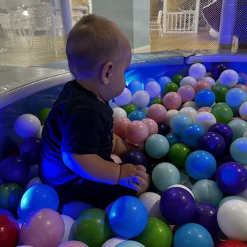 Cheeky Monkeys Fairview - 4 years & younger ball pit area! - A toddler is seated in a ball pit with multicolored plastic balls, inside an indoor children's play area with visible play structures in the background. - Playgrounds Near Me - Fairview, Texas