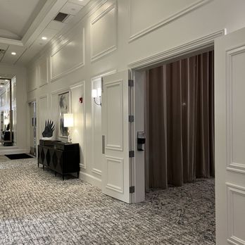 The Grandview - Newly renovated pre-function space - The image shows an interior hallway of an event venue with white paneled walls and decorative molding. A black console table with a lamp and artwork is positioned along the wall. To the right, there is an open doorway with brown curtains, possibly leading to a separate room or space. The ceiling has recessed lighting and a drop ceiling with tiles. The floor is covered with a patterned carpet, and the overall decor is modern and elegant. - Venues & Event Spaces Near Me - Poughkeepsie, New York