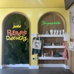 The image shows the interior of Junbi, a business specializing in matcha products. The wall is painted yellow with two arched niches; one contains a green moss wall with the text 'junbi REPRE DARINGLY,' and the other has a neon sign reading 'I love you so matcha.' Shelves hold various matcha-related items, and a tote bag with 'KYOTO' hangs on a hook. The space includes lighting fixtures and a portion of a television or monitor in the upper right corner.