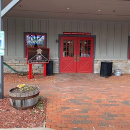 the entrance to the machine shed - The image shows the front entrance of The Wisconsin Machine Shed restaurant, featuring red double doors with glass panes. There is a sign in front of the building with the restaurant's name and logo, and a neon Bud Light sign in the window. The area has brick pavement, a wooden planter with plants, and two black trash receptacles on either side of the entrance. The building's exterior is clad in gray siding with stone accents at the base.