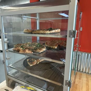 Desi Boys Pizza - A glass display case showcases multiple pizzas, including a large pizza with toppings on the top shelf and other pizzas on lower shelves. The case is positioned against a red wall with framed certificates or licenses hanging above it. The setting appears to be a restaurant or pizzeria, with a wooden floor and some paper napkins or tissues at the base of the case. - Pizza Near Me - Chicago, Illinois
