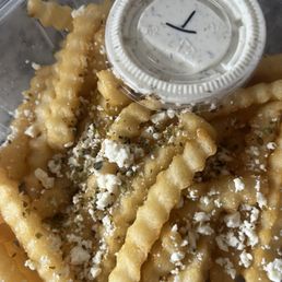 Feta Greek Friesfries - A serving of crinkle-cut French fries topped with shredded cheese, herbs, and crumbled cheese is shown in a clear container. A small plastic cup of creamy white dipping sauce is placed on top of the fries. The fries are golden and crispy, with visible seasoning and toppings, and the sauce appears to be a ranch or similar dressing.